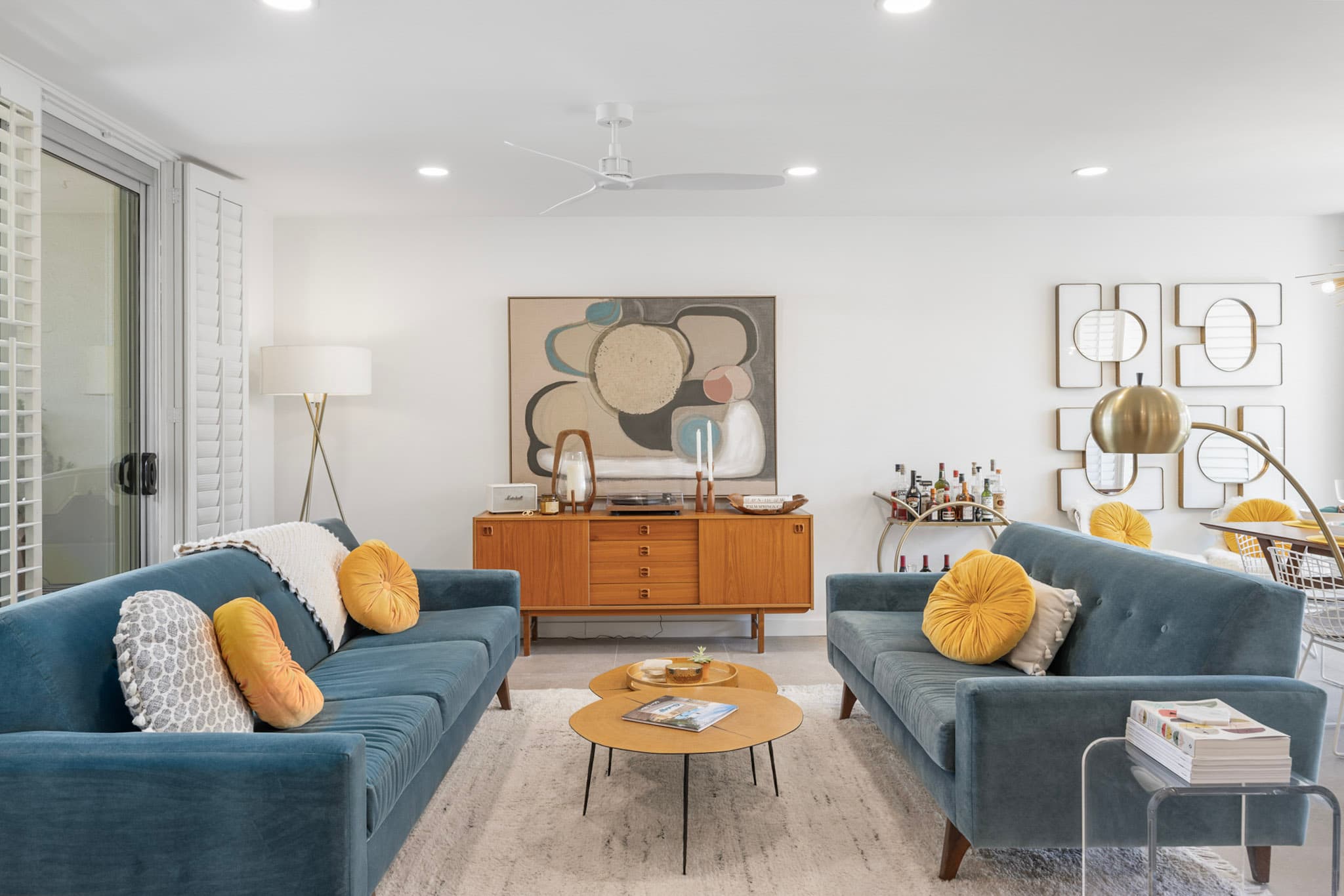 Mid-century modern living room in Palm Springs: two teal velvet sofas facing across round wood coffee tables, with a teak credenza and a large abstract canvas behind